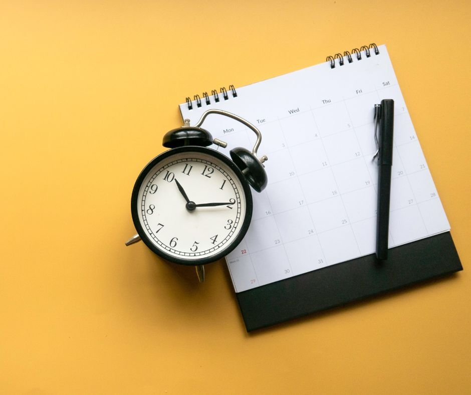 A black analog alarm clock, spiral-bound calendar, and black pen rest on a vibrant yellow background, symbolizing the organizational tools often utilized by virtual assistants to efficiently manage tasks.