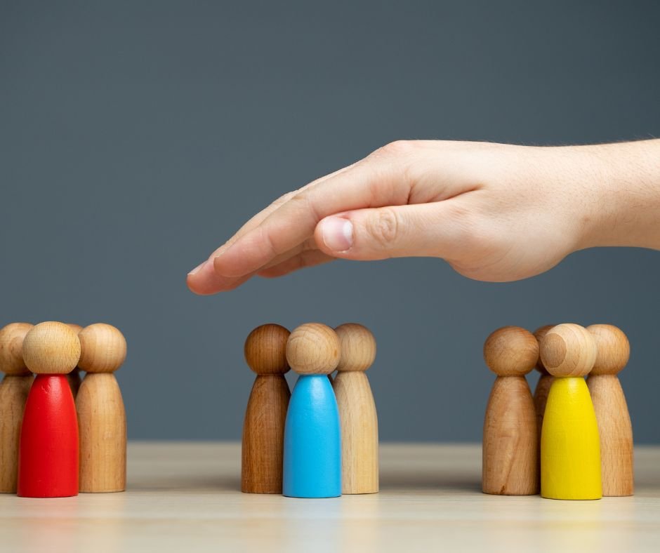 A hand hovers over groups of wooden figures on a table, with one group separated from the rest, like a remote work team managed by a virtual assistant. The figures are painted in various colors: red, blue, and yellow.