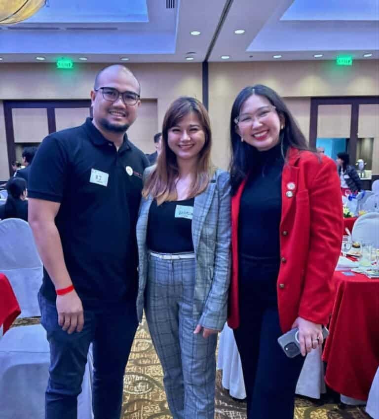 Three people stand together and smile at an indoor event with round tables and chairs in the background. Two wear business attire, and one wears a black polo shirt, representing Create and Reach VA—a virtual assistant company for cleaning services.
