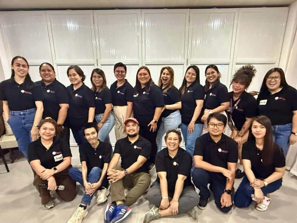 A group of sixteen people wearing matching black shirts and jeans pose and smile together indoors against a white paneled background, representing a dedicated create and reach VA team for your cleaning business virtual assistant company.