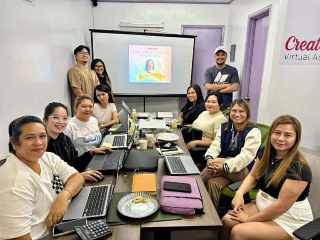 A group of people, part of the Create and Reach VA team, sit and stand around a conference table with laptops, facing the camera; a presentation is projected on the screen behind them.