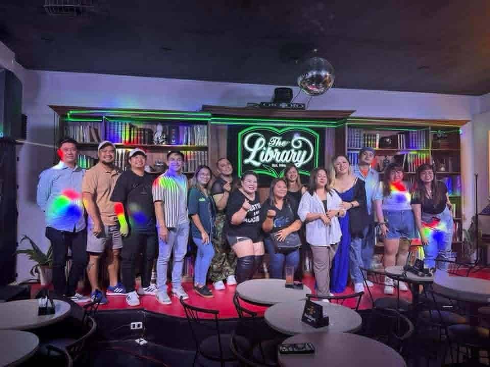 A group of people pose together on stage at The Library comedy bar, with neon signage and bookshelves in the background—celebrating the success of a leading virtual assistant company for cleaning services.
