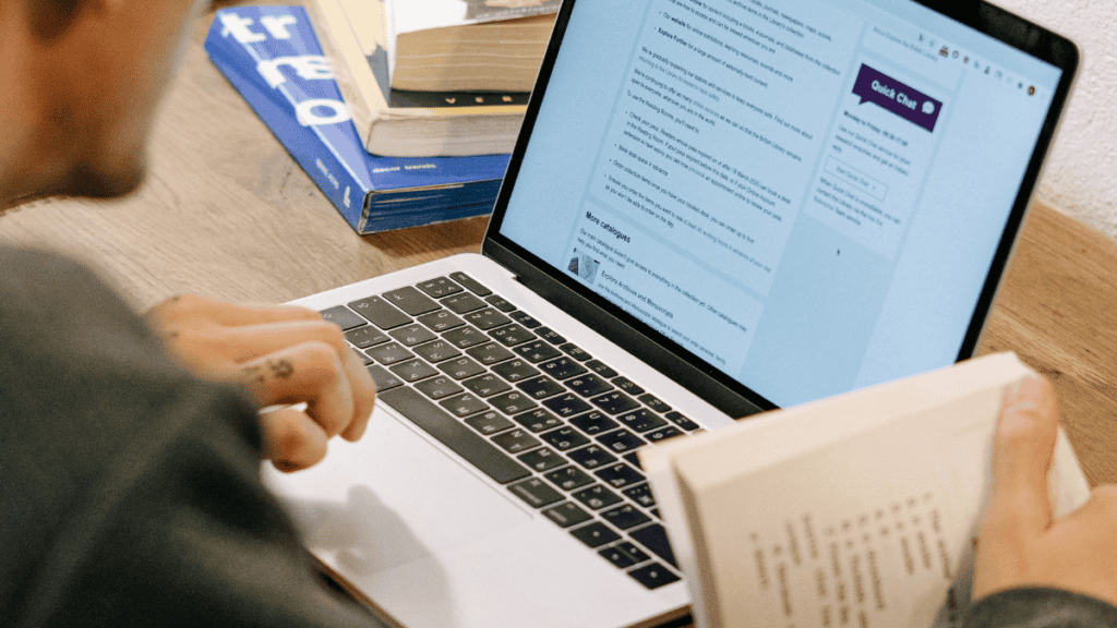 Person reading a book and looking at a laptop screen displaying text, with additional books on a wooden table.