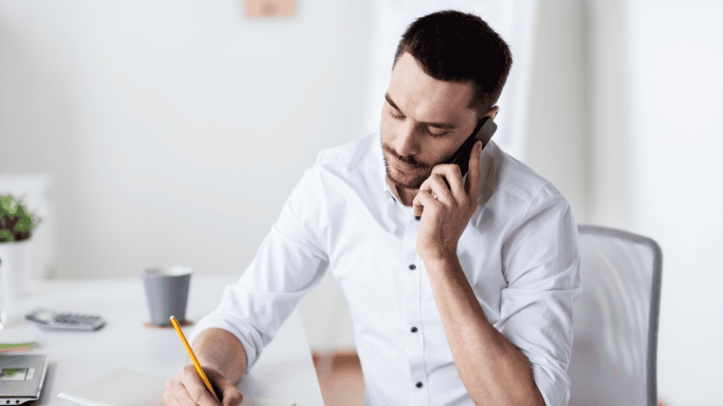 A man in a white shirt sits at a desk, talks on the phone, and writes in a notebook.