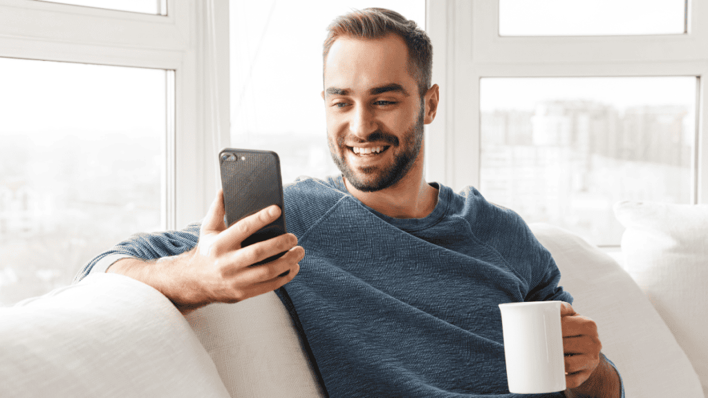 A man with a beard sits on a couch by a window, smiling at his smartphone while holding a white mug in his other hand.