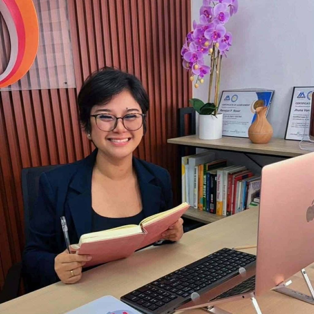 A person with short hair and glasses sits at a desk, smiling, holding an open notebook and pen—perhaps planning for a virtual assistant company for cleaning services—with a computer, books, and certificates in the background.