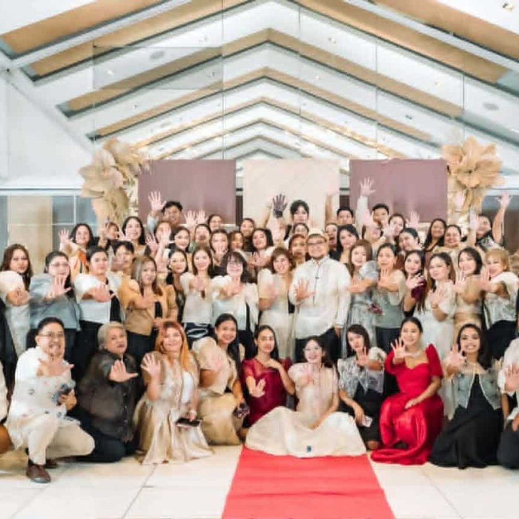 A large group of people in formal attire pose together indoors, many holding up one hand, with a red carpet and decorative backdrop behind them—representing Create and Reach VA, a leading virtual assistant company for cleaning services.