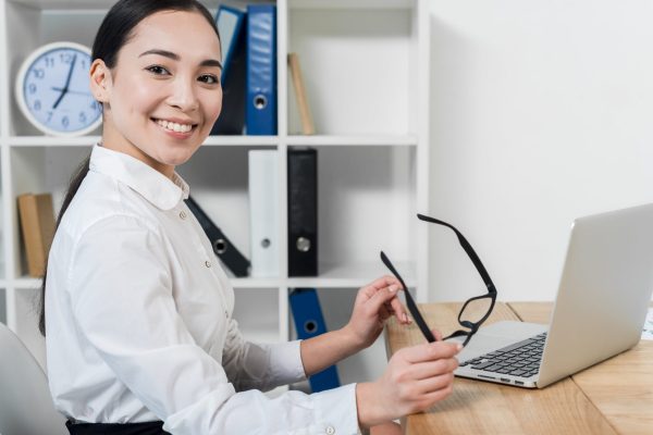 portrait-smiling-young-businesswoman-holding-eyeglasses-hand-with-laptop-desk-min-scaled